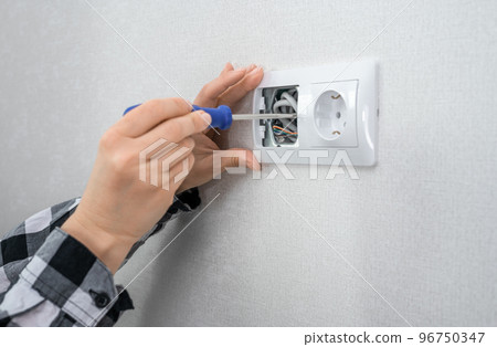 Hand with screwdriver for repairing electrical outlets at home. Electrician is using screwdriver to install power outlet in to plastic box on white wall. 96750347