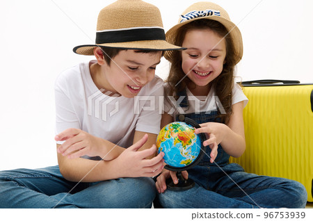 Happy children sitting near yellow suitcase, brother and sister going abroad for summer holidays, pointing fingers at place on globe with Earth map, choosing new travel destination for weekend getaway Happy children sitting near yellow suitcase, brother and sister going abroad for summer holidays, pointing fingers at place on globe with Earth map, choosing new travel destination for weekend getaway 96753939