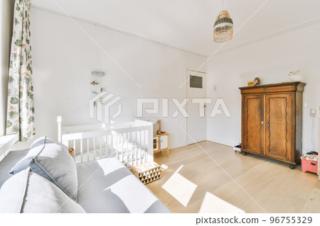 Interior of white room with baby crib and armchair near window with blue chest of drawers in daylight 96755329