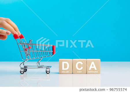 The text DCA is written on wooden cubes and woman's hand pushingn empty shopping cart in studio on blue background. Business concept. DCA - short for dollar cost averaging. 96755574