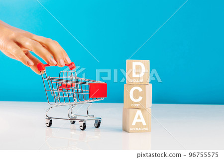 The text DCA is written on wooden cubes and woman's hand pushingn empty shopping cart in studio on blue background. Business concept. DCA - short for dollar cost averaging. 96755575