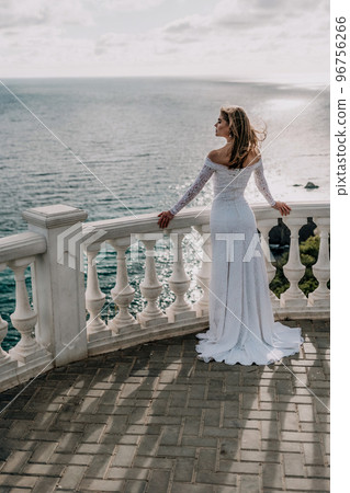 Romantic bride, blond girl in white wedding dress with open shoulders posing on open terrace with backdrop of the sea and rocks. Stylish young woman standing on terrace and looking on ocean at sunset Romantic bride, blond girl in white wedding dress with open shoulders posing on open terrace with backdrop of the sea and rocks. Stylish young woman standing on terrace and looking on ocean at sunset 96756266