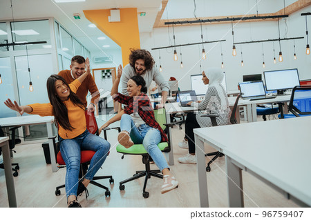 Team building and office fun. Young cheerful businesspeople in smart casual wear having fun while racing on office chairs and smiling. Team building and office fun. Young cheerful businesspeople in smart casual wear having fun while racing on office chairs and smiling. 96759407