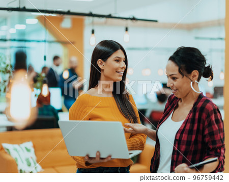 Portrait of two businesswomen talking to each other while standing in a modern business office with their colleagues using laptop and tablet.. Marketing concept. Multi-ethnic society. Portrait of two businesswomen talking to each other while standing in a modern business office with their colleagues using laptop and tablet.. Marketing concept. Multi-ethnic society. 96759442