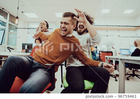 Team building and office fun. Young cheerful businesspeople in smart casual wear having fun while racing on office chairs and smiling. 96759461