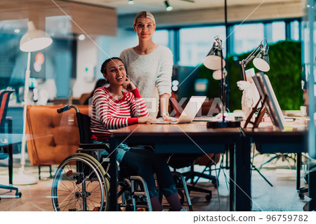 Businesswoman in a wheelchair working in a creative office. Business team in modern coworking office space. Colleagues working in the background at late night. Businesswoman in a wheelchair working in a creative office. Business team in modern coworking office space. Colleagues working in the background at late night. 96759782