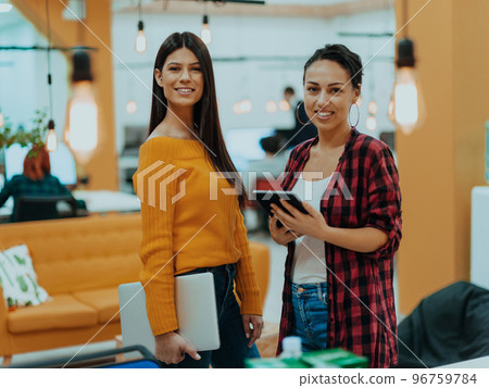 Portrait of two businesswomen talking to each other while standing in a modern business office with their colleagues using laptop and tablet.. Marketing concept. Multi-ethnic society. Portrait of two businesswomen talking to each other while standing in a modern business office with their colleagues using laptop and tablet.. Marketing concept. Multi-ethnic society. 96759784