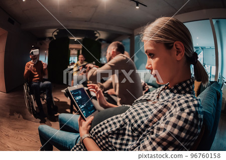 Pregnant woman sitting on the sofa of a modern office surrounded by colleagues and using a tablet 96760158