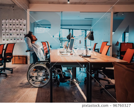 Businessman with a disability in wheelchair working overtime alone at his desk in an office late at night Businessman with a disability in wheelchair working overtime alone at his desk in an office late at night 96760327