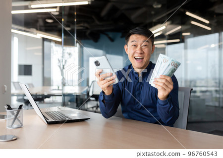 Portrait successful businessman asian smiling contentedly and looking at camera holding phone and money cash dollars in hands, celebrating victory, man in shirt working inside office using laptop. Portrait successful businessman asian smiling contentedly and looking at camera holding phone and money cash dollars in hands, celebrating victory, man in shirt working inside office using laptop. 96760543