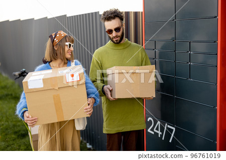 Man and woman carry parcels received in automatic post machine outdoors 96761019