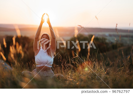 Young Woman Sitting in Meditation Yoga Pose and Catching Sun by Hands at Sunset Young Woman Sitting in Meditation Yoga Pose and Catching Sun by Hands at Sunset 96762797