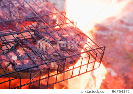 Cooking chicken meat over charcoal. Chicken meat fried with a crust on a wire rack during grill cooking. Backlight from a burning fire and smoke in the late evening. 96764371