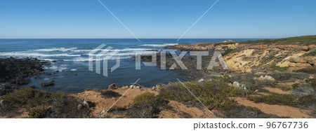 Panoramic view of sea shore, wild Rota Vicentina coast with ocean waves, sharp rock and green and red leaves of sour fig flower near Vila Nova de Milfontes, Portugal. Sunny day, blue sky. 96767736
