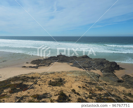 View of Praia dos Aivados sand beach with ocean waves and sharp rock and distant figures at wild Rota Vicentina coast near Porto Covo, Portugal. View of Praia dos Aivados sand beach with ocean waves and sharp rock and distant figures at wild Rota Vicentina coast near Porto Covo, Portugal. 96767751