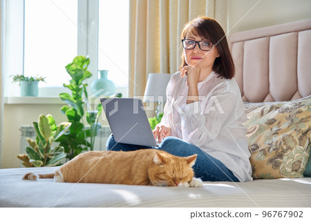 Middle aged woman at home on couch with laptop and cat 96767902