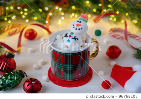 Preparing for Christmas. A red plaid mug with Snowman and marshmallows on a background of Christmas decorations and fir branches with an illuminated garland. Winter Christmas scene. Preparing for Christmas. A red plaid mug with Snowman and marshmallows on a background of Christmas decorations and fir branches with an illuminated garland. Winter Christmas scene. 96768105