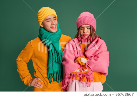Portrait of young man and woman in bright knitted hat and scarf posing isolated over green background. Making faces Portrait of young man and woman in bright knitted hat and scarf posing isolated over green background. Making faces 96768667