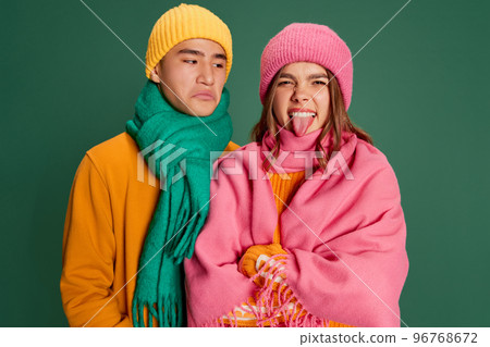 Portrait of young man and woman in bright knitted hat and scarf posing isolated over green background. Grimace Portrait of young man and woman in bright knitted hat and scarf posing isolated over green background. Grimace 96768672