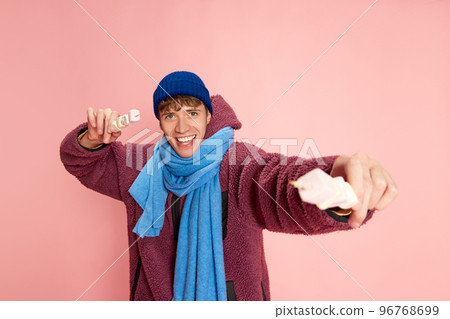 Portrait of happy smiling young man in winter clothes posing with marshmallow isolated over pink background Portrait of happy smiling young man in winter clothes posing with marshmallow isolated over pink background 96768699