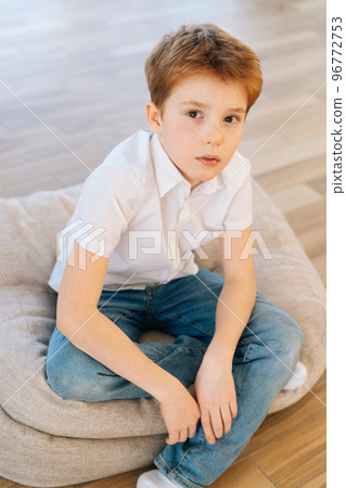 Vertical portrait of handsome child boy looking at camera sitting on pillow on floor in living room with light modern interior. Top view of serious kid posing at home alone, top view. Vertical portrait of handsome child boy looking at camera sitting on pillow on floor in living room with light modern interior. Top view of serious kid posing at home alone, top view. 96772753