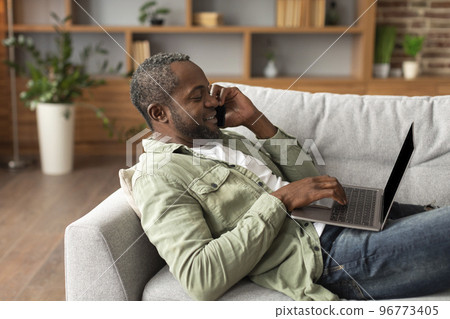 Glad busy middle aged african american male typing on laptop, calling by smartphone, choosing order Glad busy middle aged african american male typing on laptop, calling by smartphone, choosing order 96773405