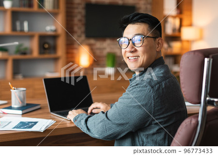 Smiling middle aged asian businessman in glasses works on computer with blank screen, sits at table in office 96773433