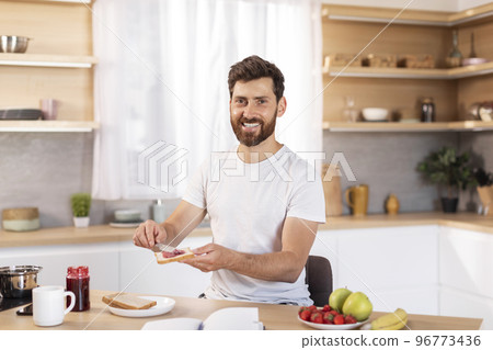 Cheerful middle aged caucasian male with beard in white t-shirt makes sandwich with fruits for breakfast 96773436