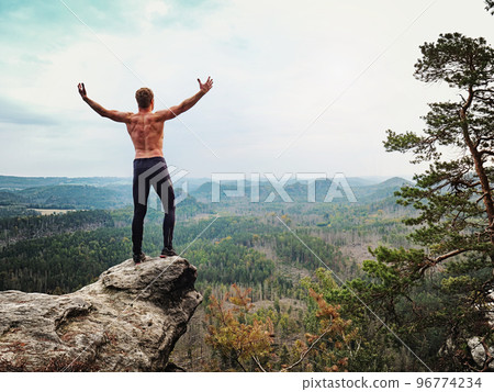 Happy shirtless man shouting with raised arms to the landscape bellow from sharp rock Happy shirtless man shouting with raised arms to the landscape bellow from sharp rock 96774234