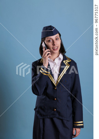 Smiling flight attendant talking on smartphone, holding mobile phone, having conversation. Stewardess in uniform answering call, air hostess standing with telephone in hand, looking at camera 96775317