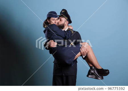 Smiling captain holding stewardess in arms portrait, aircrew in professional uniform looking at camera. Happy aviator and flight attendant romantic couple studio medium shot 96775392