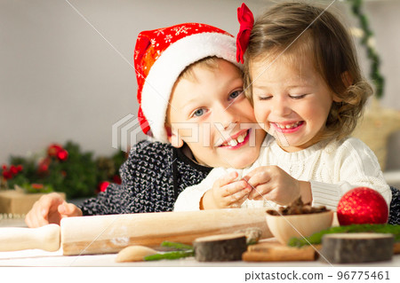 Little cute girl 2-4 with a red bow and boy 7-10 in a Christmas cap make gingerbread cookies in the New Year's kitchen. Little cute girl 2-4 with a red bow and boy 7-10 in a Christmas cap make gingerbread cookies in the New Year's kitchen. 96775461