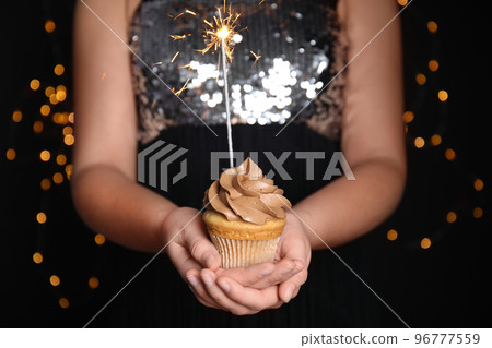 Woman holding birthday cupcake with sparkler on blurred background, closeup Woman holding birthday cupcake with sparkler on blurred background, closeup 96777559