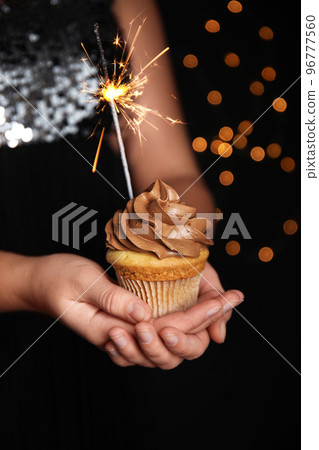 Woman holding birthday cupcake with sparkler on blurred background, closeup Woman holding birthday cupcake with sparkler on blurred background, closeup 96777560