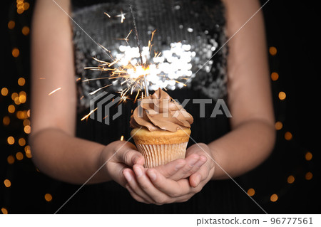 Woman holding birthday cupcake with sparkler on blurred background, closeup Woman holding birthday cupcake with sparkler on blurred background, closeup 96777561