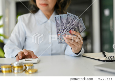 A successful and wealth Asian aged female holding US dollar bills and using calculator. cropped A successful and wealth Asian aged female holding US dollar bills and using calculator. cropped 96779553