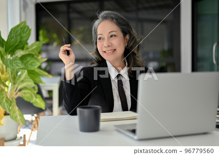 Charming Asian senior female manager sitting at her desk, smiling, daydreaming about her success. Charming Asian senior female manager sitting at her desk, smiling, daydreaming about her success. 96779560
