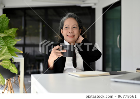 Happy Asian senior businesswoman sipping coffee and daydreaming while sitting at her desk. Happy Asian senior businesswoman sipping coffee and daydreaming while sitting at her desk. 96779561
