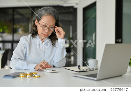 Professional Asian senior female accountant touching her eyeglasses, managing project budget 96779647