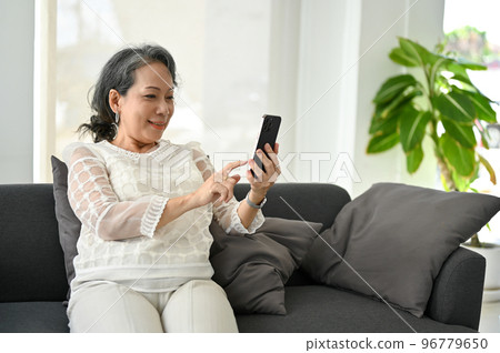 Happy Asian retire woman using her smartphone, chatting with her grandchild while relaxing on sofa Happy Asian retire woman using her smartphone, chatting with her grandchild while relaxing on sofa 96779650
