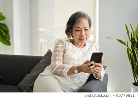 Happy Asian senior woman using her smartphone while relaxing on sofa in the living room. Happy Asian senior woman using her smartphone while relaxing on sofa in the living room. 96779651