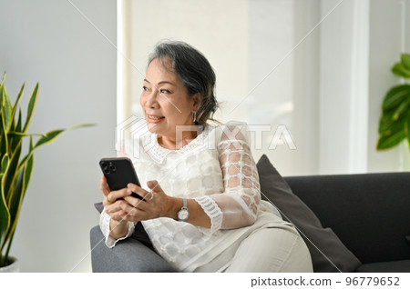 Happy Asian senior woman using her smartphone, looking at the window and daydreaming 96779652