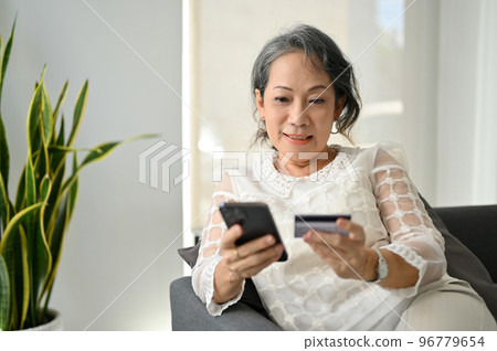 Asian aged woman using her smartphone and credit card, enjoys online shopping at home. Asian aged woman using her smartphone and credit card, enjoys online shopping at home. 96779654