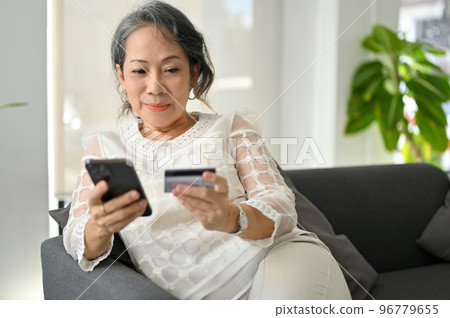 Relaxed Asian aged retire woman sitting on sofa, holding a credit card and her smartphone. Relaxed Asian aged retire woman sitting on sofa, holding a credit card and her smartphone. 96779655