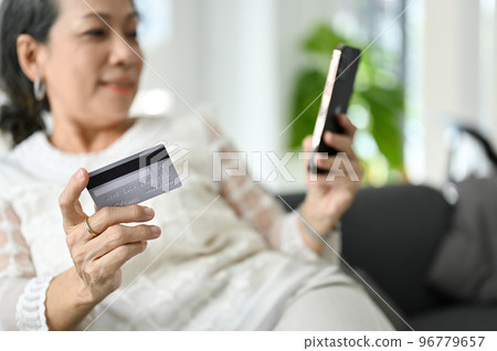 Asian aged woman relaxing on her sofa and using her smartphone to shopping online. Asian aged woman relaxing on her sofa and using her smartphone to shopping online. 96779657