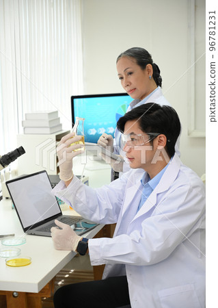 A talented young Asian male scientist in a white coat and goggles in the lab while his coworker A talented young Asian male scientist in a white coat and goggles in the lab while his coworker 96781231