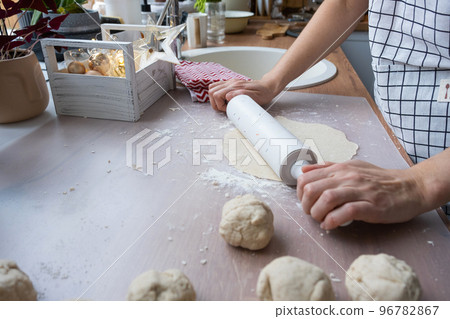 Hands knead thick dough, roll out with a rolling pin on the kitchen table, decorated with festive decorations for Christmas and New year. Baking at home, aroma and comfort. Hands knead thick dough, roll out with a rolling pin on the kitchen table, decorated with festive decorations for Christmas and New year. Baking at home, aroma and comfort. 96782867