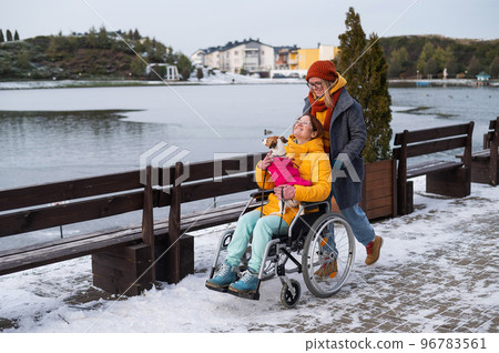 A woman in a wheelchair walks with her friend and a dog by the lake in winter. 96783561
