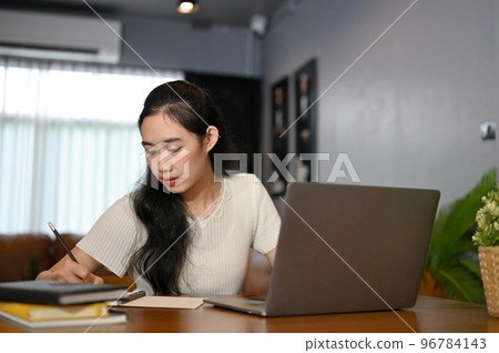 Smart young Asian female college student concentrating doing her homework in living room Smart young Asian female college student concentrating doing her homework in living room 96784143