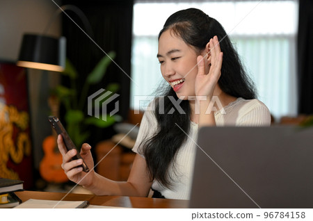 Cheerful Asian female waving hand to say Hi, video call with her friends on smartphone. Cheerful Asian female waving hand to say Hi, video call with her friends on smartphone. 96784158
